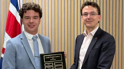 Government of Jersey Two people standing indoors against a light wooden panelled wall. One person is holding a dark plaque with gold lettering that reads “Colin Powell Award” and a list of engraved name plates beneath. A second person beside them is helping to present the award.