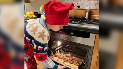 Dan Harris Joshie Harris pictured putting a rack of pies in the oven in his kitchen. He is wearing a red hat and a multi-coloured jumper with gingerbread men on and red trousers with Santa on.
 