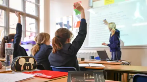 PA Media A teacher with blonde hair is writing on a whiteboard at the front of a classroom. There are three teenage girls sitting with their backs to the camera. Two of them are raising their hands to answer a question. There are pencil cases, folders and a copy of Romeo and Juliet sitting on the desk in the foreground. 