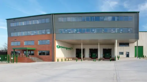 A large building with 'gompels' on the side. Part of the building juts out over the entrance, held up by white support struts. The rest of the building has large windows and walls of red brick