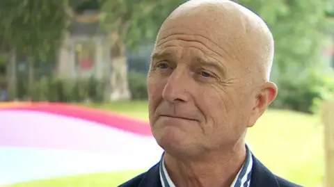 A screengrab of Tony Gribben during a BBC interview.  He has a shaved head and is wearing a dark jacket and a stripped blue and white shirt. 