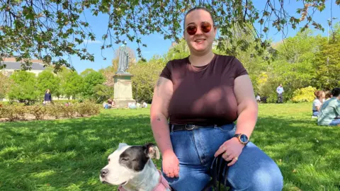 BBC A young woman wearing a brown t shirt and jeans kneeling next to a black and white Staffordshire Bull Terrier in a pink harness in a sunny park.
