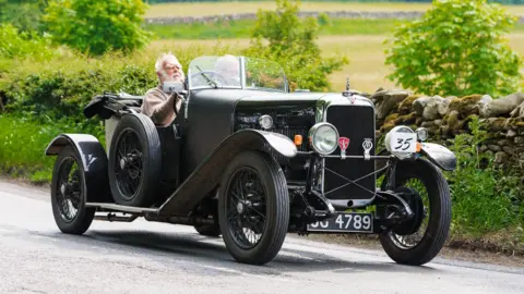 Dave Ranson A very old open-top car is driven along a country lane by a man with a white beard.