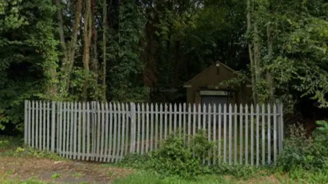 Google A single-storey brick building set back in woodland, with a metal security fence in the foreground 