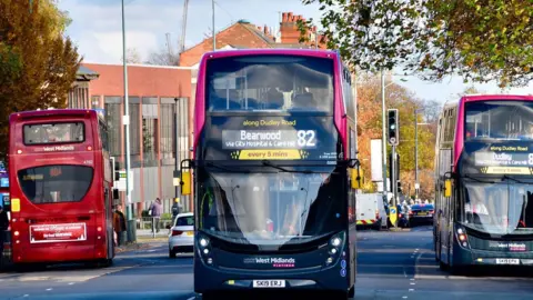 A three-lane road with three buses on it. A red bus is at a bus stop facing away from the camera. Another bus, with Dudley marked as its destination, is at a bus stop on the other side of the road facing the camera. Between them, a bus heading for Bearwood is driving towards the camera.