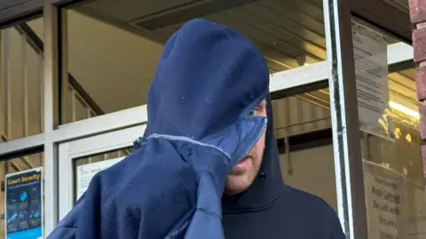 A close-up image of a man wearing a black hooded top with the hood of a navy coat pulled over his head and part of his face as he tried to evade the camera.