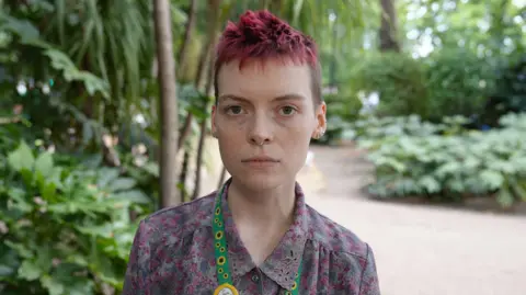 Qays Najm/BBC A person with short spiky hair looking at the camera. They are wearing a floral shirt and a green lanyard with sunflowers on it. Behind them is a path and greenery.