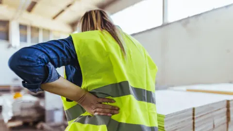 Getty Images A stock image of a woman in a hi-vis yellow vest holding on to her back in pain in a warehouse environment. 