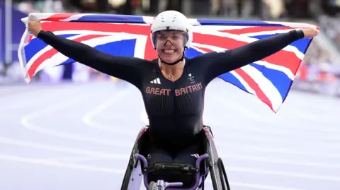 PA Media Hannah Cockroft holding the Union Jack up after a race. She is sat in her wheelchair and wears a dark navy Team GB skinsuit and helmet. The flag is hoisted behind her head. She is smiling.