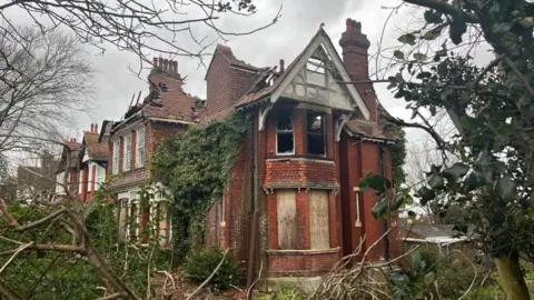 BBC/George Carden A derelict red brick house with boarded up windows which has been ravaged by fire.
