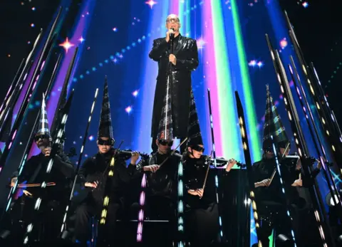 Getty Images Neil Tennant standing on a platform in the middle of rays of multi-coloured lights, above a row of violinists wearing tall pointy black hats