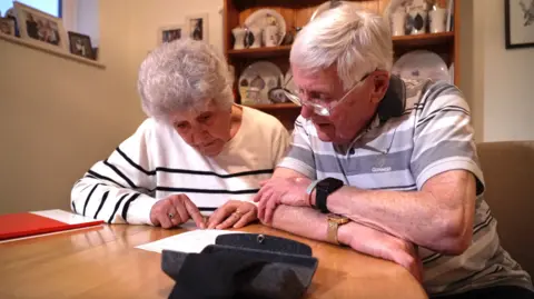 Maureen and Eric, both 85, are looking over the invoice they were given. They are sitting at a table together reading the invoice