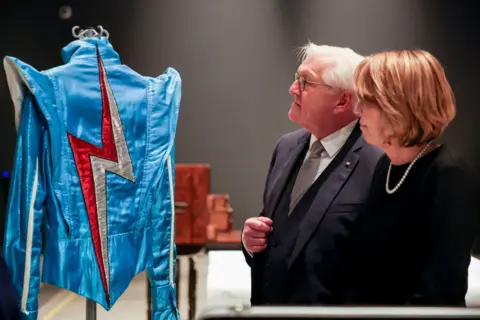 Steinmeier and Budenbender walk near a jacket on display at a museum. The jacket is bright blue with dramatic shoulder pads and a large red and silver lighting bolt covering the length of its back.