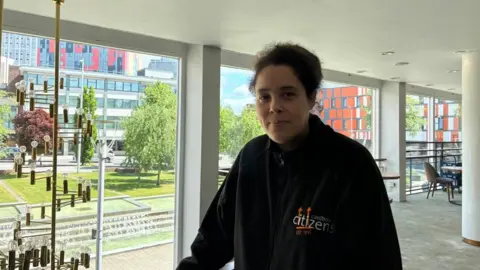 A black woman stands in front of big glass windows and wearing a black hoodie.