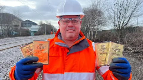 Rob Cochrane is standing by a railway line holding four video cassettes in his hands. The boxes are gold with black lettering. He is wearing an orange hi-vis jacket, a white safety helmet and blue gloves.