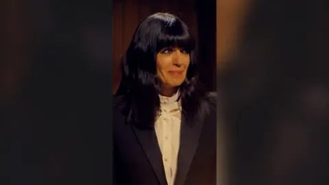 Claudia Winkleman looks to the side, smiling. She has long black hair and wears a white shirt and a black blazer