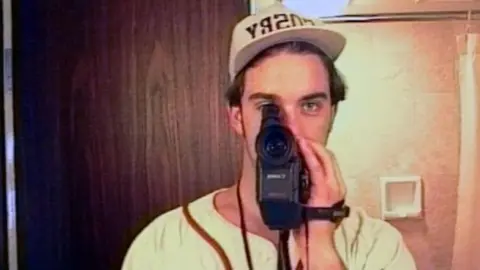 Take That/Netflix Robbie Williams, who is wearing a white baseball cap and jersey with red detailing, is holding a camcorder up to his right eye.