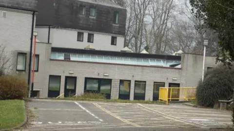 Facebook A general view of the assessment centre, which is grey brick with a row of ground floor windows and a row of skylight windows above. A concrete car park can be seen in front of the building with a yellow railing on the right hand side.