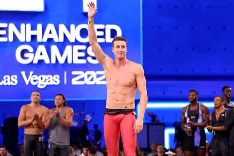 An athlete stands with right arm raised, wearing skintight pink spandex trousers.