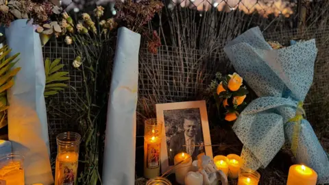 A small black and white picture of John George is placed in the grass next to a wire fence. Candles and flowers bring it to the fence and the surrounding grass to light. John is dressed in the photo and he is smiling. Beyond the fence, a small amount of orange light fills the night sky. 