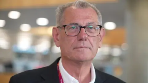Zoie O'Brien/BBC A head and shoulders shot of a man with short white hair, wearing black-framed glasses and a dark jacket over a white shirt. He is inside.
