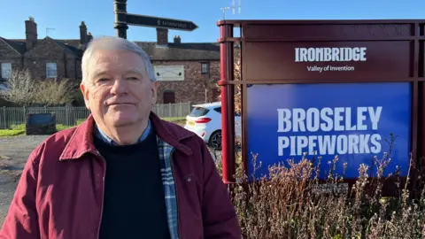 A man with grey hair, a burgundy jacket, navy jumper and blue shirt. To his left is a sign reading "Ironbridge Valley of Invention: Broseley Pipeworks."
Behind him you can see the museum itself - a brick building the size of a house, with a large beige sign outside with a symbol of two crossed tobacco pipes.
The sky is blue and clear, and the weather is sunny casting shadows on the ground. 