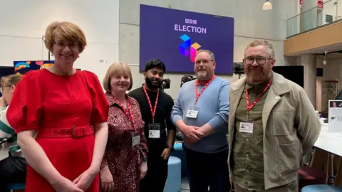 Two women and three men stand in front of a screen showing a BBC Election logo.