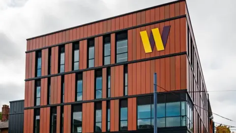 A three storey brown building with wide windows and a yellow W sign in the top right corner.