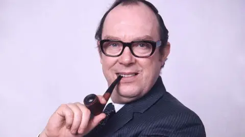 Head and shoulders image of Eric Morecambe smoking a pipe in a dark blue striped suit in 1973