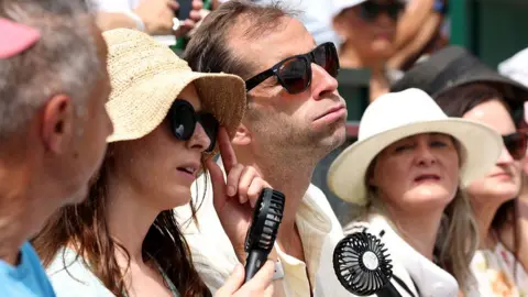 A row of people sat. The picture shows only their heads. A man in the middle looks like he is puffing in the heat, as he uses a hand fan and wears sun glasses. The women on either side wear wide straw hats, one wears sunglasses and also has a fan.