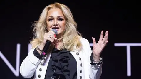 Bonnie Tyler performs in Madrid on 28 September 2024. She has a white jacket and shoulder-length light-coloured hair and she is singing into a microphone on a dark stage.