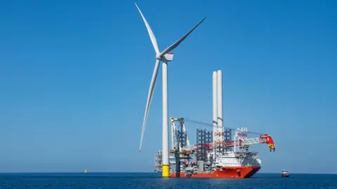 A wind turbine in the sea. Next to it, there is a large ship being used in the turbine construction process. The blue sea is flat and calm. The cloudless sky is blue.