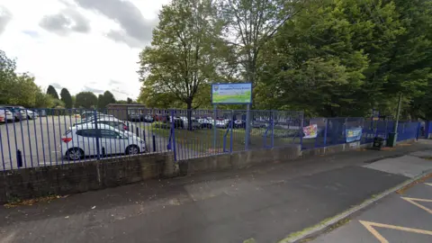 Google The car park of a school which has concrete for parking, cars lines up but also a grass area and trees. There is a blue metal fence surrounding it. Beyond the cars is the school building 