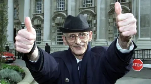 PA Media Man wearing dark navy overcoat and black hat stands with two thumbs up to the camera outside Belfast High Court