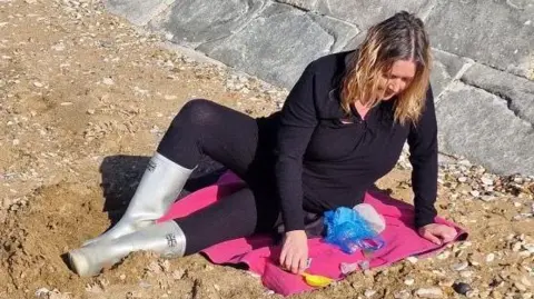 Angela Schrier on the beach at Hunstanton. She is wearing an all black outfit with silver wellies. She is sitting on the beach on a pink towel.