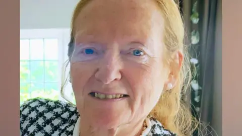 A woman in her late 60s, with blue eyes and light ginger hair tied back, smiles at the camera. She is inside a house with a green curtain and window behind her, and is wearing a black and white chequered jacket. 