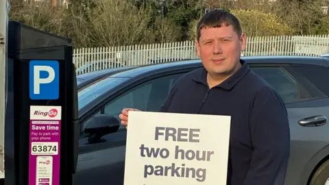 A man holds a white board with the words 'free two hour parking'. He is stood in a car park, with a pay and display machine to his right. A car can be seen behind him.