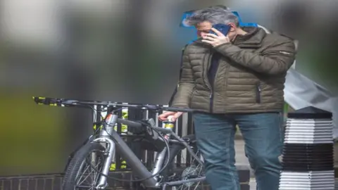 Met Police Howard Phillips wearing a brown coat and jeans while on the phone. He is using his left hand to press a USB stick into the seat of a bicycle.