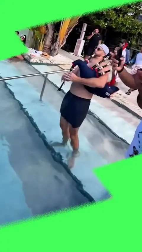 Man in shorts playing bagpipes in front of a pool, with palm trees and hotel behind.