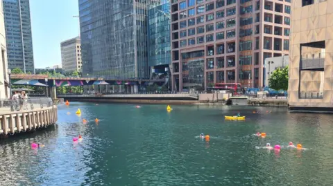 Love Open Water Swimmers in water at Canary Wharf with tall glass-fronted buildings in the background along with a bridge