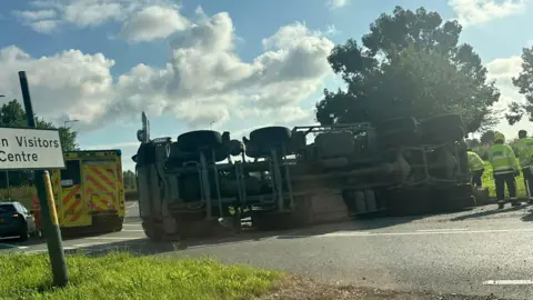 A cement mixer lorry lies on its side with its under-section towards the camera. Three firefighters stand at the rear of the vehicle, while behind it is an ambulance and a car. 