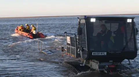 Wells RNLI An RNLI inshore lifeboat is on the left going out into the river. On the right, in the foreground, there is a vehicle which is helping to send the lifeboat out to shore.