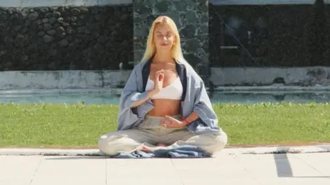 Anastasia sits cross-legged meditating in a garden on a sunny day