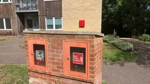 Owen Sennitt/BBC A fire hydrant box can be seen in front of a block of flats behind it, with the words "dry rise outlet" written on posters behind a screen
