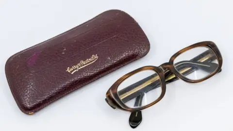 Hansons Auctioneers A maroon leather glasses case on the left and to the right is a pair of horn rimmed heavy glasses, folded up on their arms