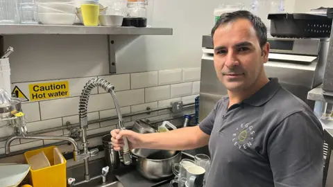 A man in a grey shirt standing in a kitchen.