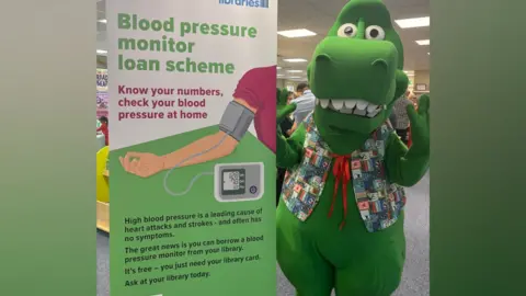 A dinosaur mascot standing next to a banner promoting blood pressure monitors. It reads: "Blood pressure monitor loan scheme. Know your numbers, check your blood pressure at home. High blood pressure is a leading cause of heart attacks and strokes - and often has no symptoms. The great news is you can borrow a blood pressure monitor from your library. It's free - you just need your library card. Ask at your library today."
