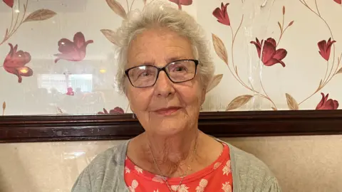 An elderly lady sits on a bench in a social club, there is a wooden rail and patterned wallpaper behind her. She is wearing a pink t-shirt, grey cardigan and glasses. She has short white hair.  