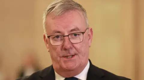 A man with grey hair and glasses looks at the camera. His facial expression indicates he is in the middle of speaking. He is wearing a dark suit although only the tops of his shoulders are visible