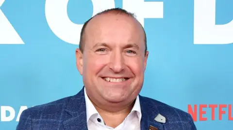 Dave Fishwick smiles as he poses at press photocall in front of a blue backdrop promoting the Netflix movie Bank of Dave. He's wearing a blue jacket over a white shirt.
